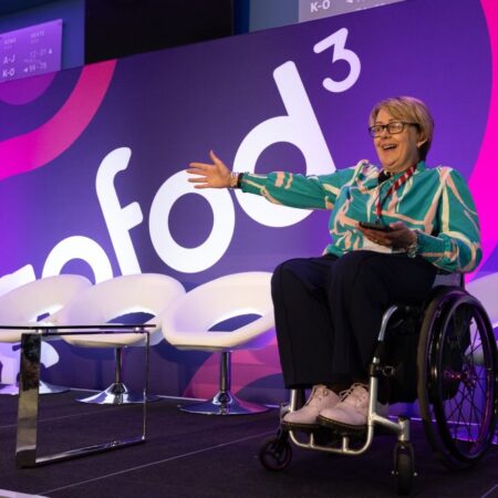 Speaker on stage in wheelchair with event logo backdrop, holding notes.