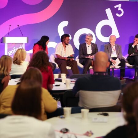 Presenter addressing audience in front of a large purple event backdrop.