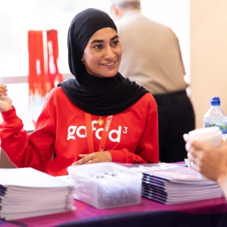 Event volunteer in a red shirt with logo assisting at an information desk.
