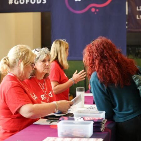 Three people in red “#gofod3” t-shirts seated at a table assisting another participant with papers or materials.