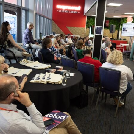 Group of attendees seated around tables listening to a speaker in a conference room.