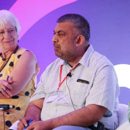 Person seated on stage wearing an event lanyard, purple backdrop behind.